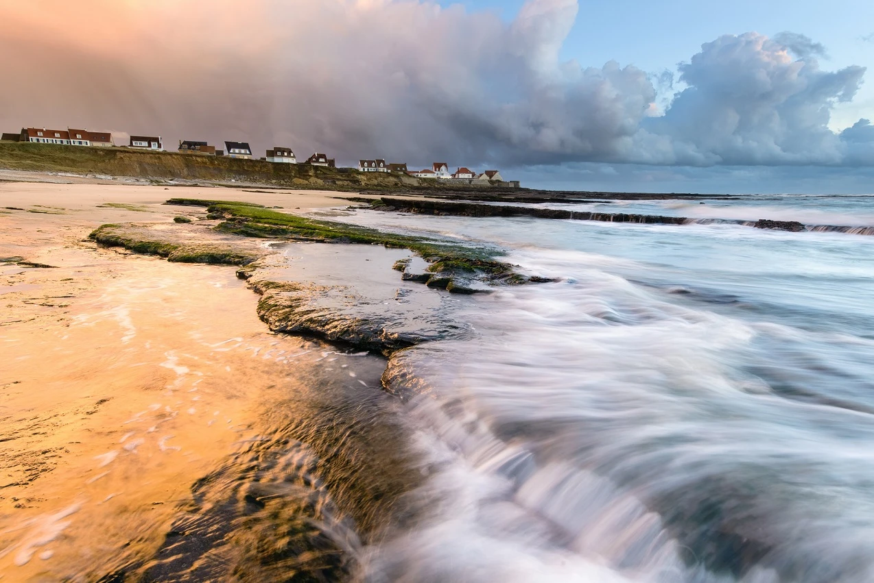 a deserted beach at sunset, in the background are houses and in the sky are cloud mountains towering out.