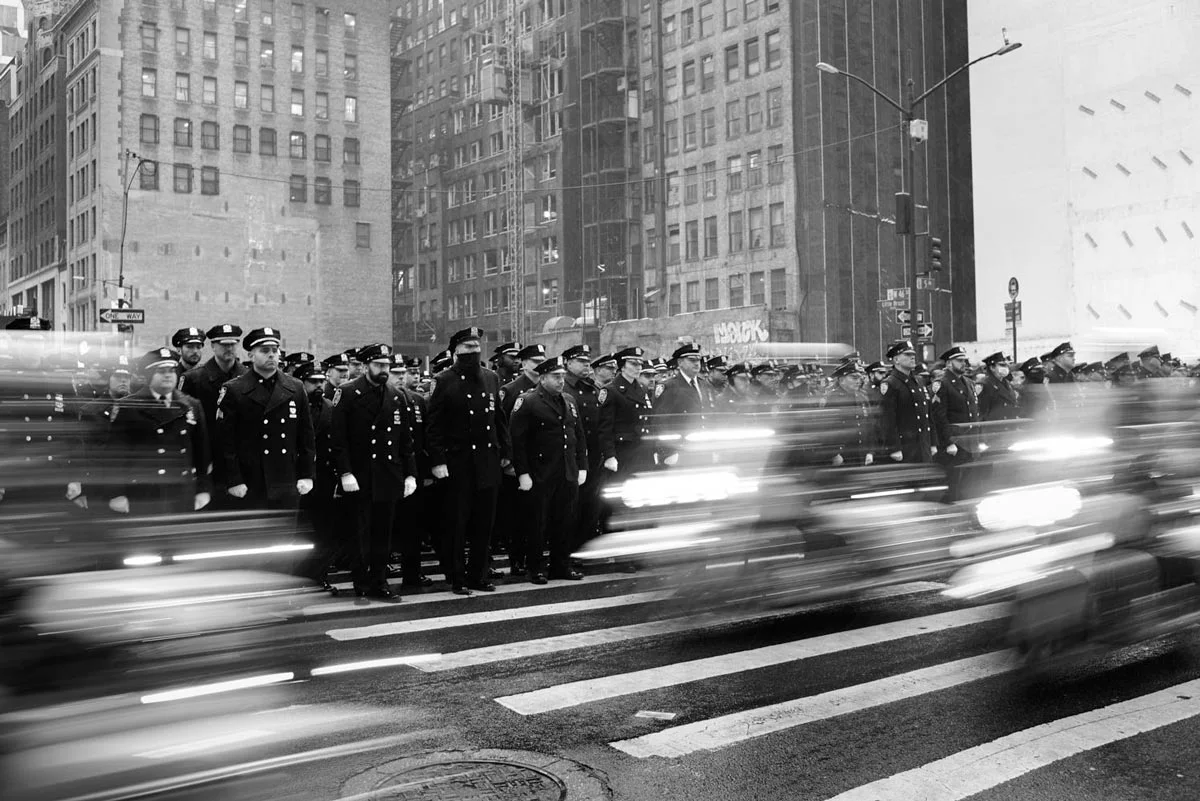 Policemen cross an intersection, motorcycles drive past, only vaguely recognizable. Image in black and white.