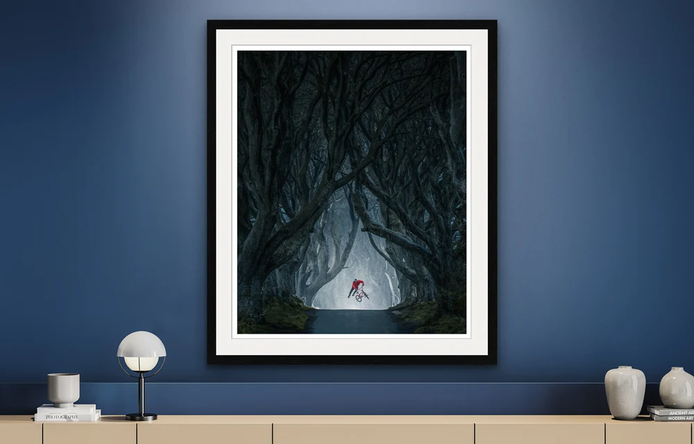 BMX rider showing a jump trick on a road in a dark forest as a Hahnemühle Fine Art Print in a passe-partout frame.