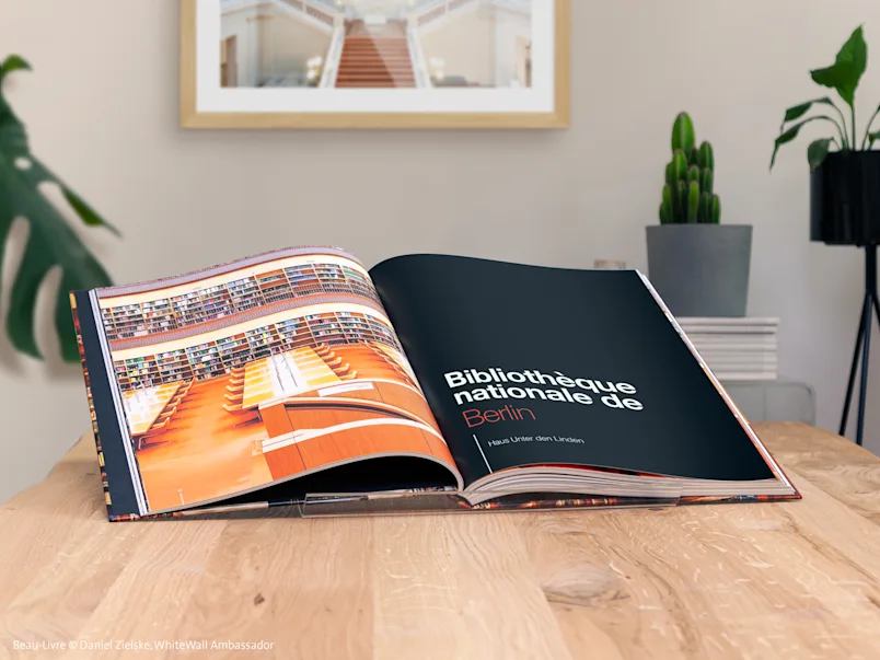 Album photo ouvert sur une table en bois, avec des photos de la Bibliothèque d'État de Berlin ; plantes et cadre photo en arrière-plan.