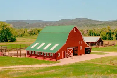 A red farmhouse with a green gabled roof in a meadow set against wooded hills.