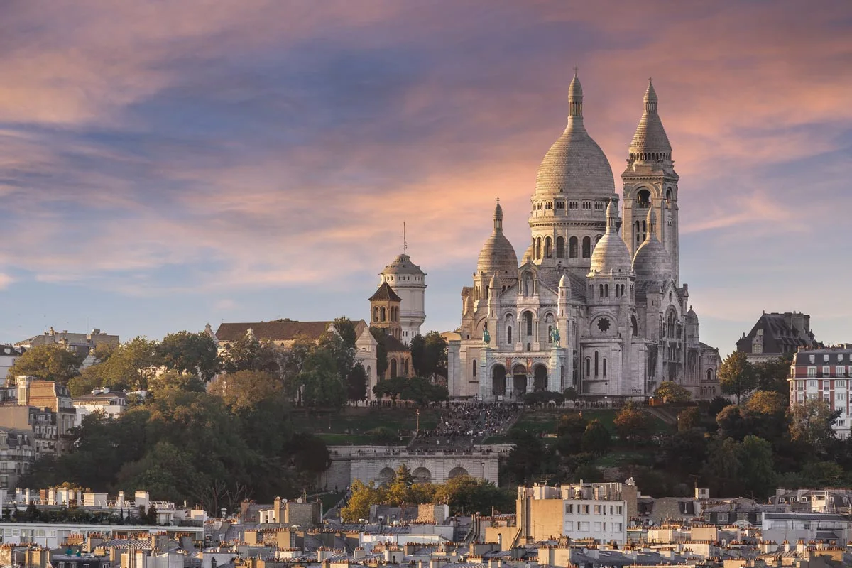 A large, white cathedral with several domes and towers stands on a hill surrounded by trees and buildings.