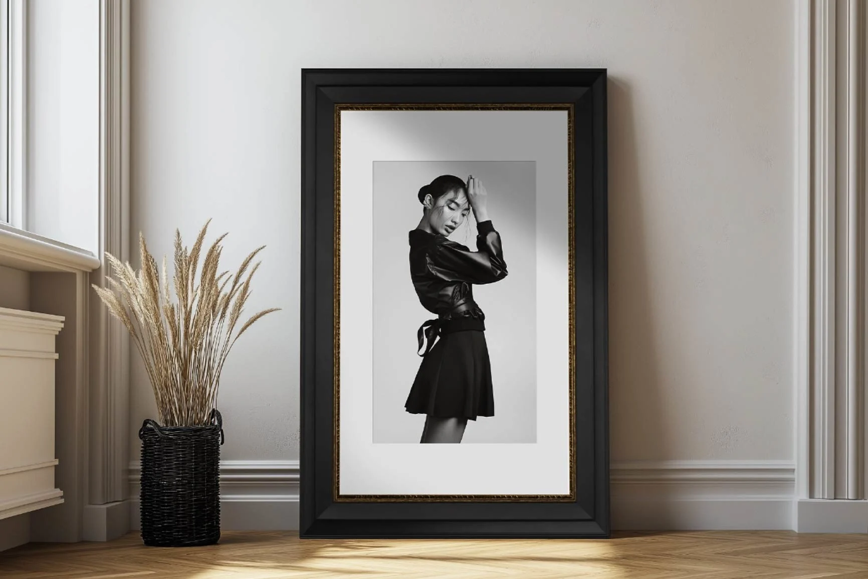Fotografia in bianco e nero di una donna in posa di profilo con giacca di pelle e gonna, incorniciata e in piedi sul pavimento davanti a una parete.