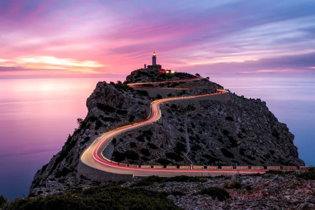 Une route sinueuse mène à un phare sur une colline rocheuse, sous un ciel dans les tons roses et bleus de Stephan Wiesner.