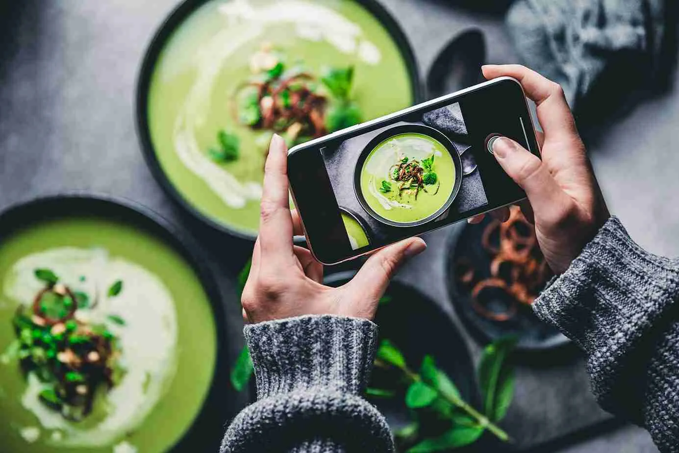 Hände mit grauem Pullover halten Smartphone und fotografieren grüne Suppe in Schale, eine weitere gefüllte Schale daneben.