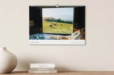 Calendar page with a window view of a green field with sheep, underneath an unmade bed, bright room.