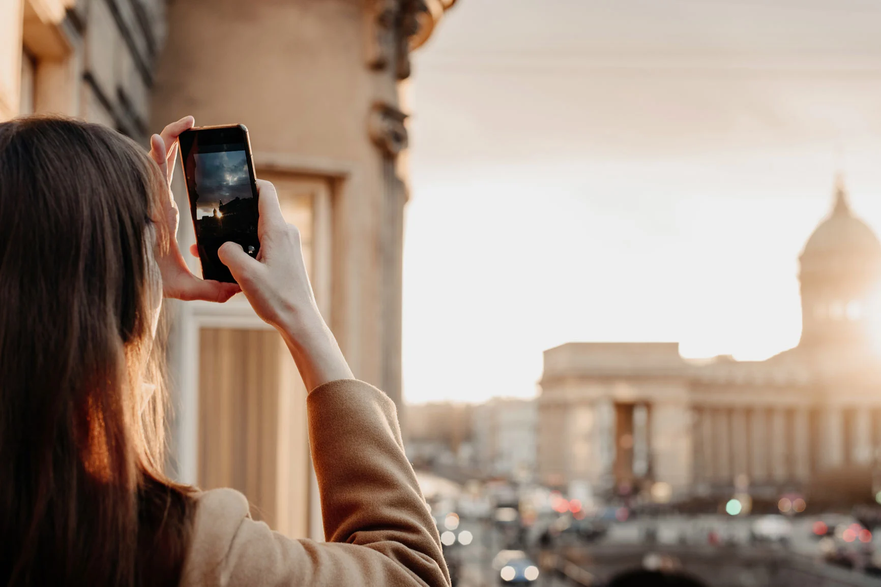 Mujer con abrigo marrón fotografiando con un smartphone una vista de la ciudad al atardecer.