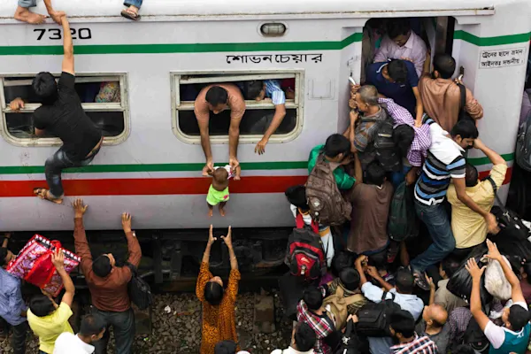A dramatic scene of a crowded train. People cling to the outside while a baby is being handed down through a window to a woman on the platform.