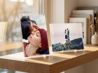 Twee acryl fotostandaards met romantische foto's op een houten plank, verlicht door zacht natuurlijk licht, met boeken en een vaas op de achtergrond.