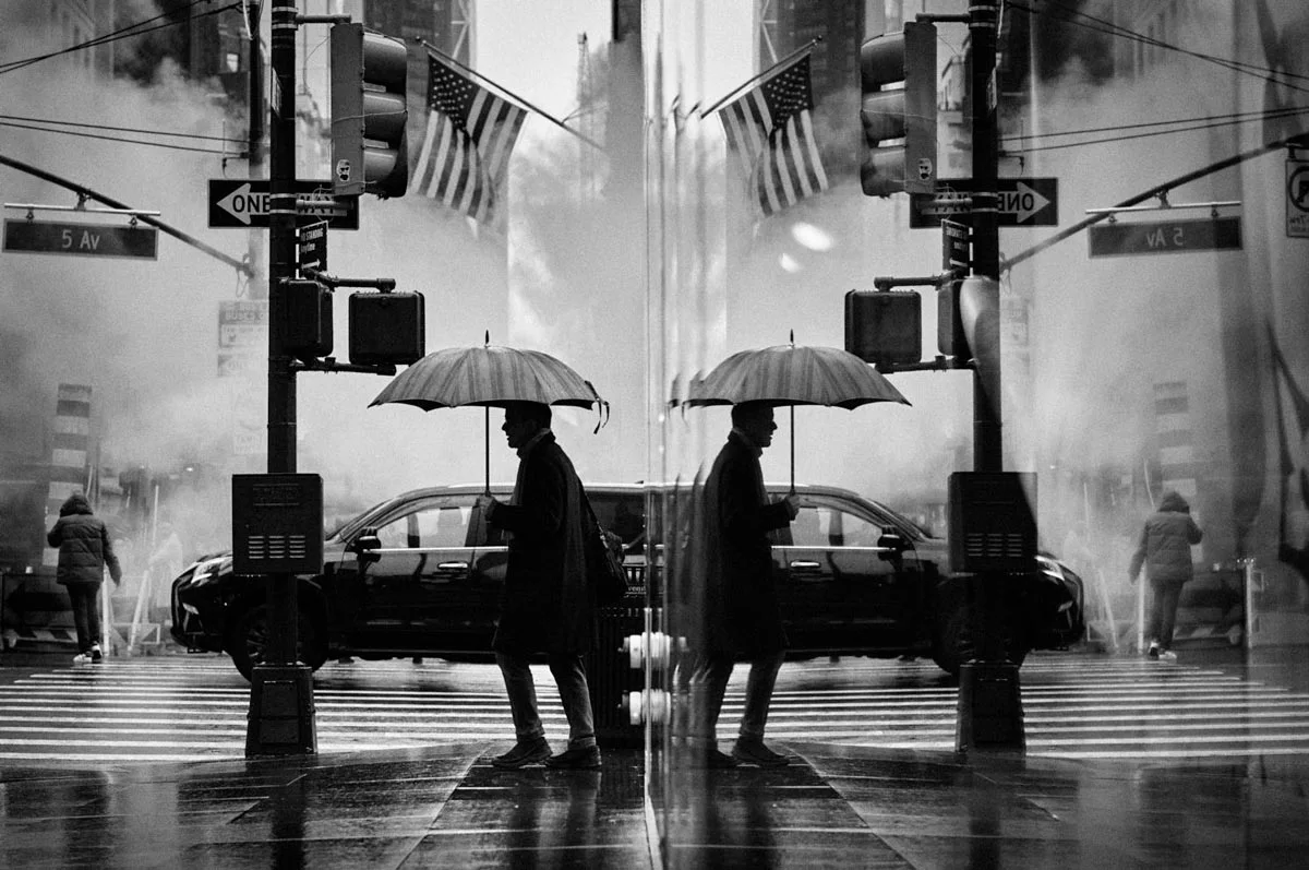 In a black-and-white photograph, a man with an umbrella stands on a sidewalk in front of an intersection while his reflection can be seen on a glass surface.