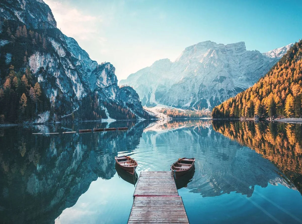 Wooden jetty with two rowboats on a tranquil mountain lake, surrounded by wooded and rocky mountains in autumn.