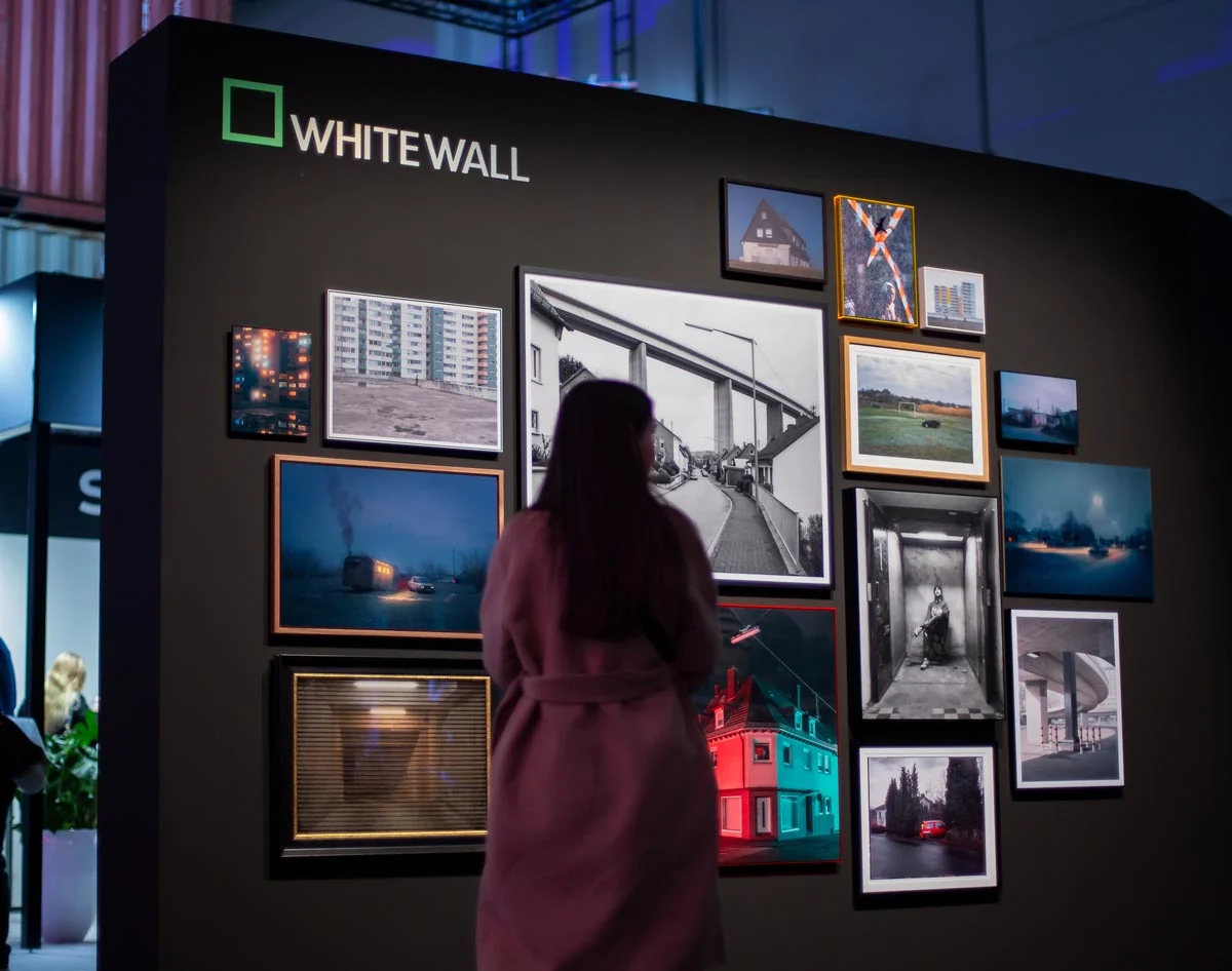 A person stands in front of a black exhibition wall with a WhiteWall logo. Several framed pictures in different sizes and styles are arranged on the wall.