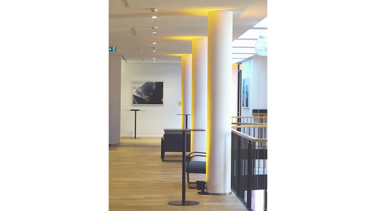 Modern lobby interior with white columns, warm yellow accent lighting, wood floors, dark furniture, and framed artwork on white walls.