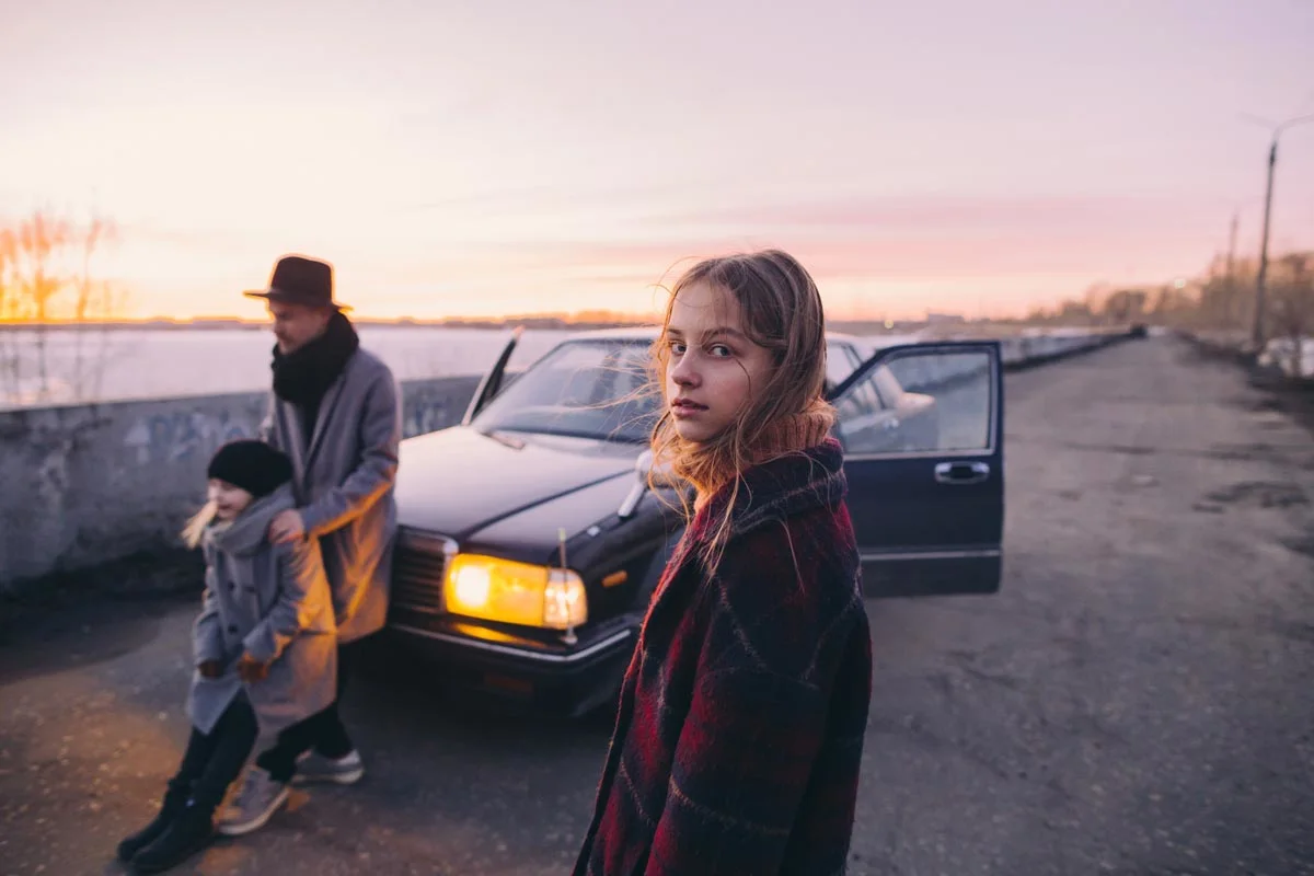 A girl stands in front of a car with open doors, in the background a man with a hat and a child standing next to the car. The sky is colored slightly pink.