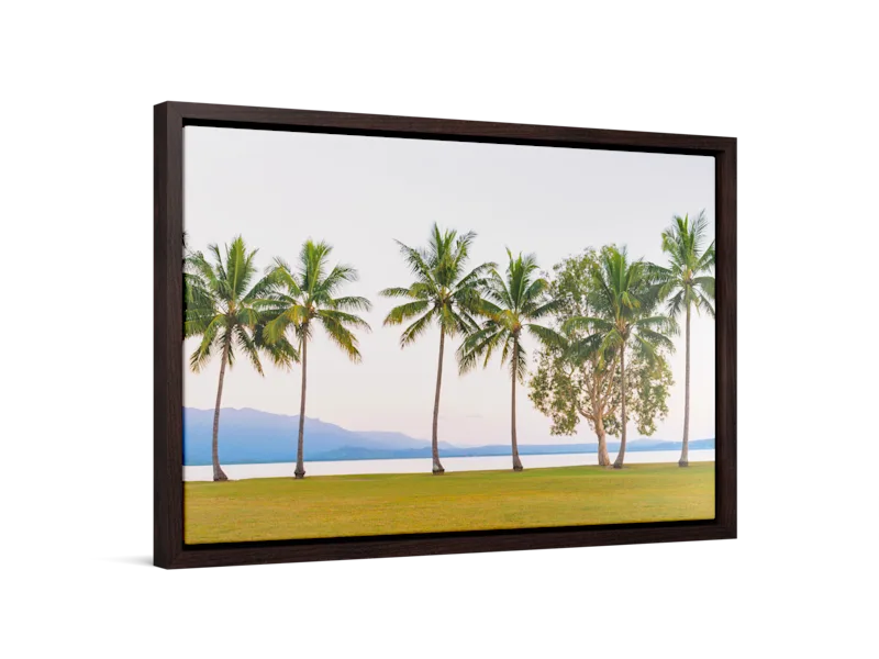 Impression sur toile encadrée représentant de grands palmiers sur une pelouse verte au bord d’une eau calme, avec au loin des montagnes bleues et un ciel pastel doux.