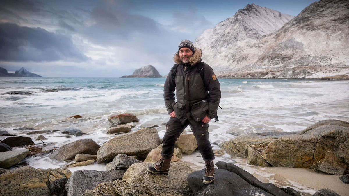 A man in a dark winter jacket and wool hat stands on rocky shore with crashing waves and snow-covered mountains behind him.