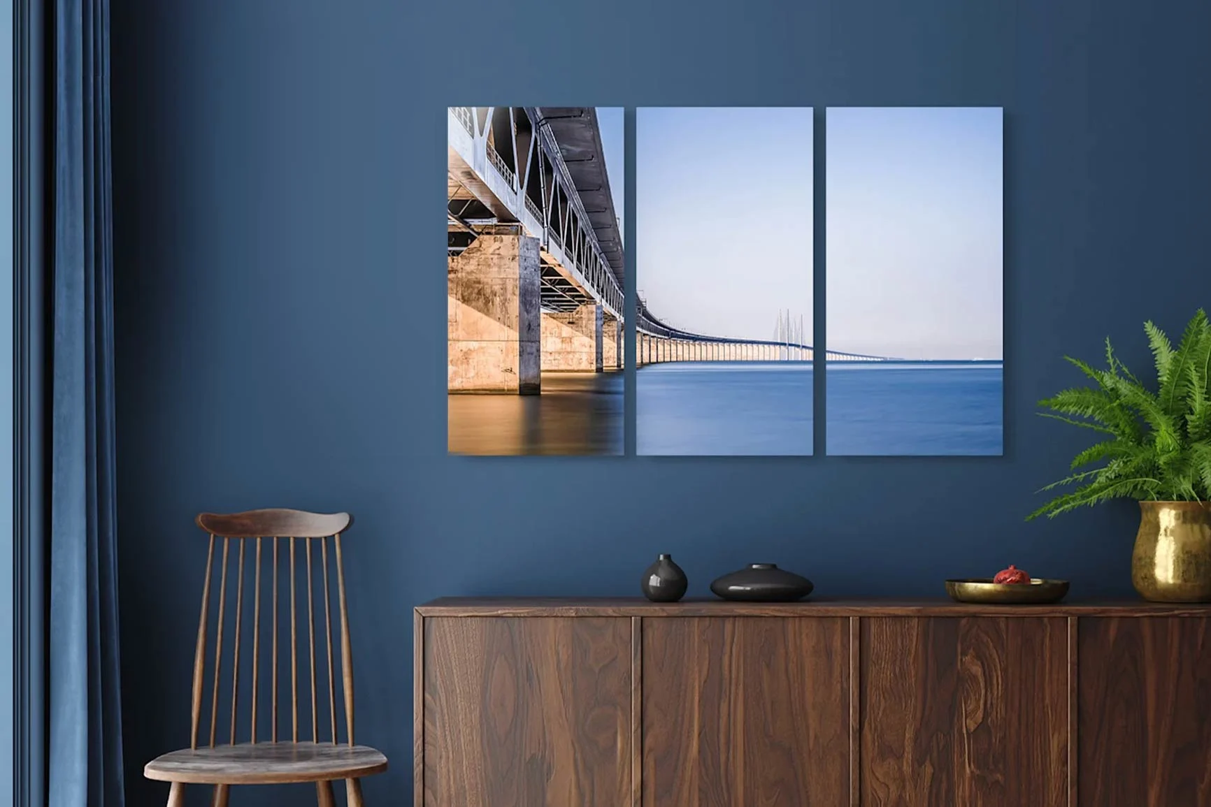 Trittico con motivo di ponti su acque tranquille e cielo pallido appeso alla parete sopra una credenza decorata e una sedia di legno.