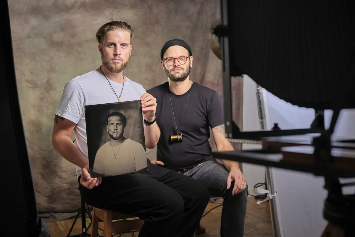 two men sitting in front of the camera, one of them holding a print of himself.