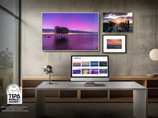 Een moderne, minimalistische werkplek met een desktopmonitor waarop de Shopify-storefront van fotograaf James Katsipis te zien is. Aan de betonnen muur erboven hangen drie hoogwaardige wanddecoraties: een groot kustmotief in levendig paars en twee kleinere zonsondergangscènes. Linksonder prijkt het zegel van de "TIPA World Awards 2026" voor de beste Shopify-integratie.