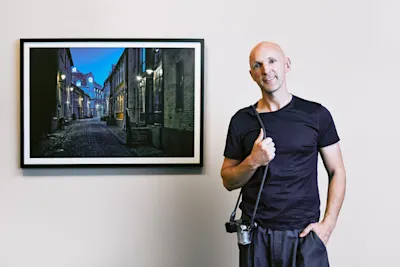 Phil Penman with camera next to a framed photo of an illuminated alleyway at dusk on the wall.