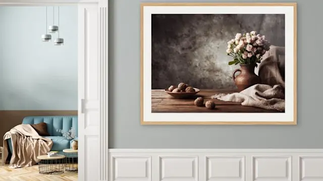 Framed still life with flowers in a vase, nuts on a wooden table and a cloth, on a gray wall.