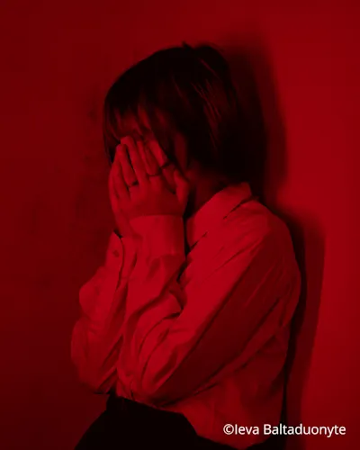 woman leaning against a dark red wall, covering her face with both hands
