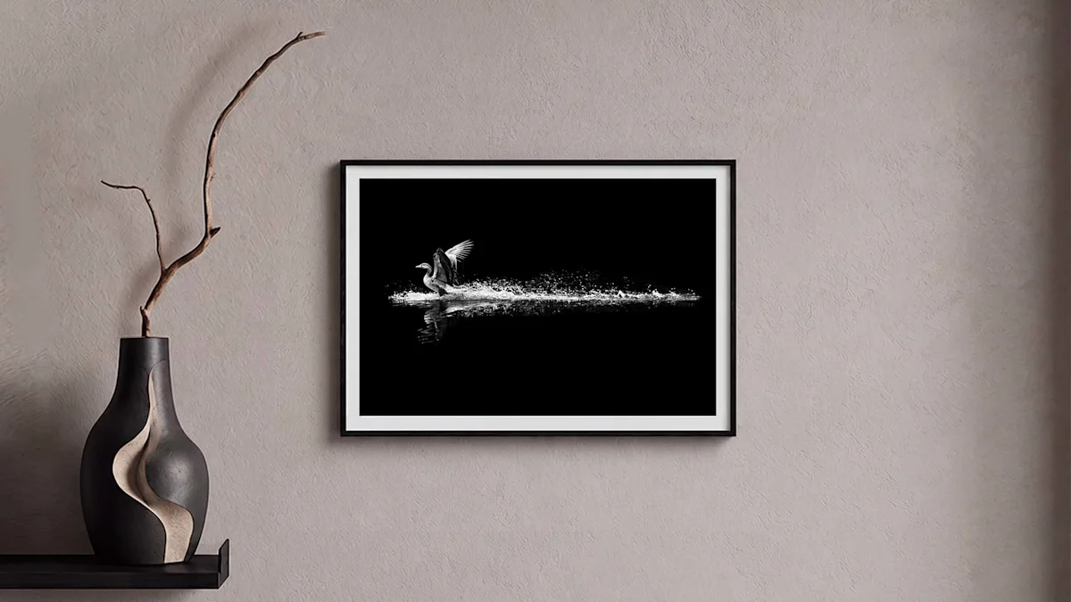 A framed black and white photo of a bird taking flight over water hangs on a wall beside a dark decorative vase.