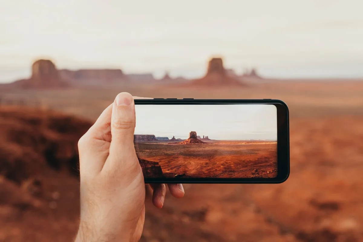 Hand hält Smartphone, zeigt Wüstenlandschaft mit Felsen und rotem Boden, unscharfer Hintergrund
