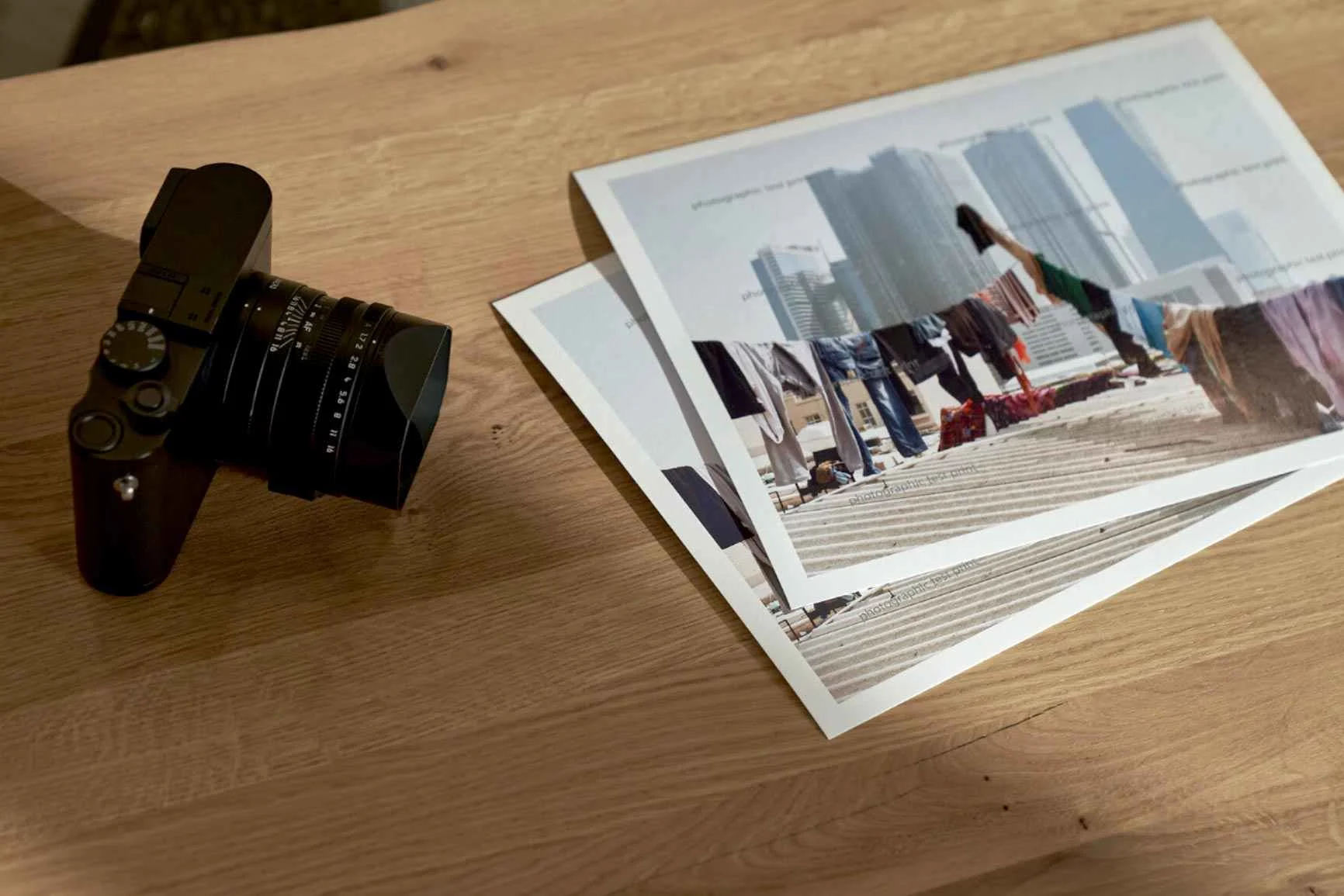 Schwarze Kamera mit Objektiv neben mehreren Fine Art Prints auf hellem Holztisch.