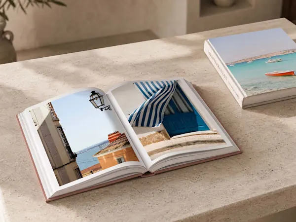 Toma desde un ángulo elevado de un libro de fotografía premium abierto con tapa dura de color salmón, apoyado sobre una superficie de piedra de travertino claro. La página izquierda exhibe una fotografía vibrante de varios tomates rojos y verdes esparcidos sobre un fondo azul claro. La página derecha presenta una toma vertical de una fachada blanca mediterránea clásica, adornada con buganvillas de color rosa brillante junto a una contraventana azul. En la esquina superior derecha del fondo, se aprecia parcialmente otro libro cerrado con una escena costera.