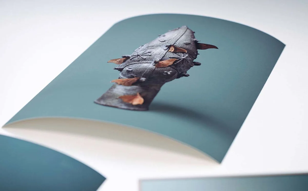 Die Fotografie zeigt einen ungerahmten Fine Art Print, der leicht gewölbt auf einer hellen Unterlage liegt. Auf dem matten Papier ist ein verziertes Bronzeobjekt mit braunen Faltern vor einem blaugrünen Hintergrund aus einer schrägen Perspektive abgebildet. Im Vorder- und Hintergrund sind Anschnitte weiterer Drucke angedeutet, während der Fokus scharf auf dem zentralen Motiv liegt. Die Aufnahme hebt die hochwertige Papierqualität und die matte Oberflächenstruktur des Drucks hervor.