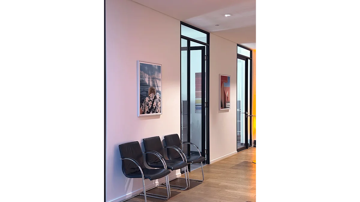 A modern office hallway with three black leather chairs, framed artwork on white walls, and black-framed glass partition doors.