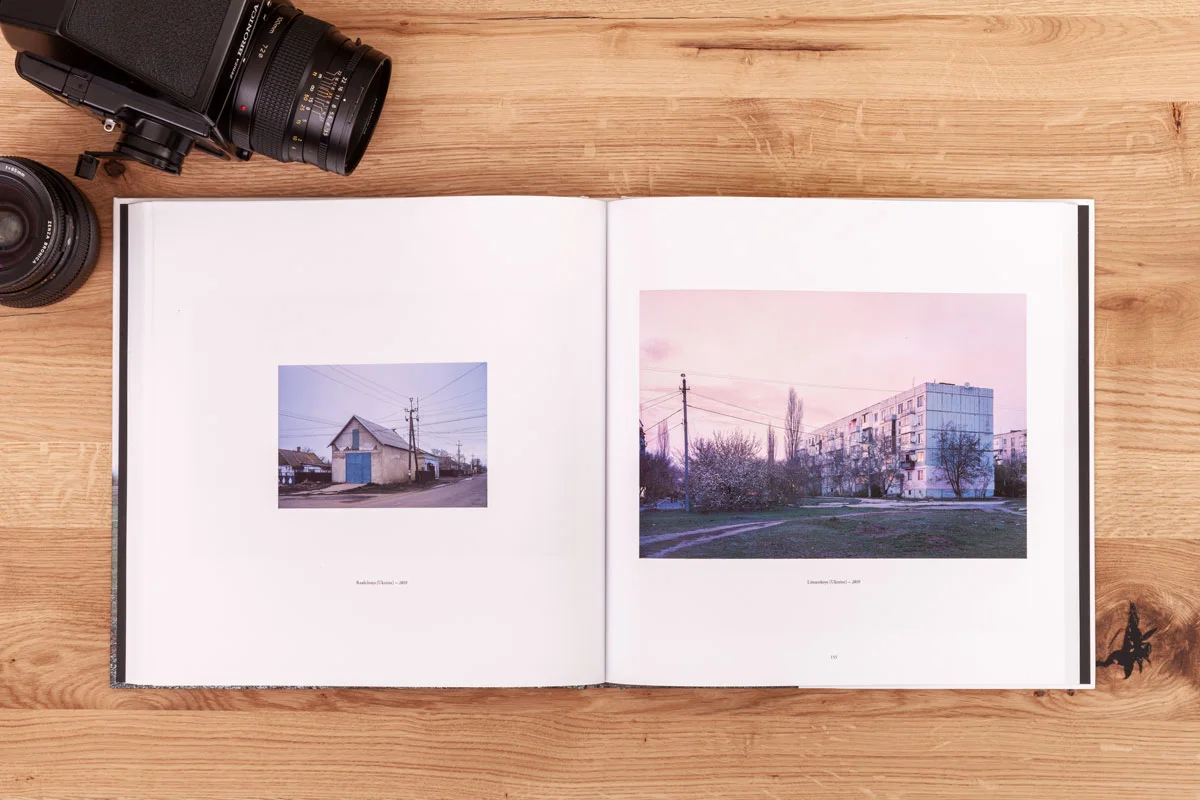 Open photo book with two pictures on a wooden surface, next to it a camera and a lens.