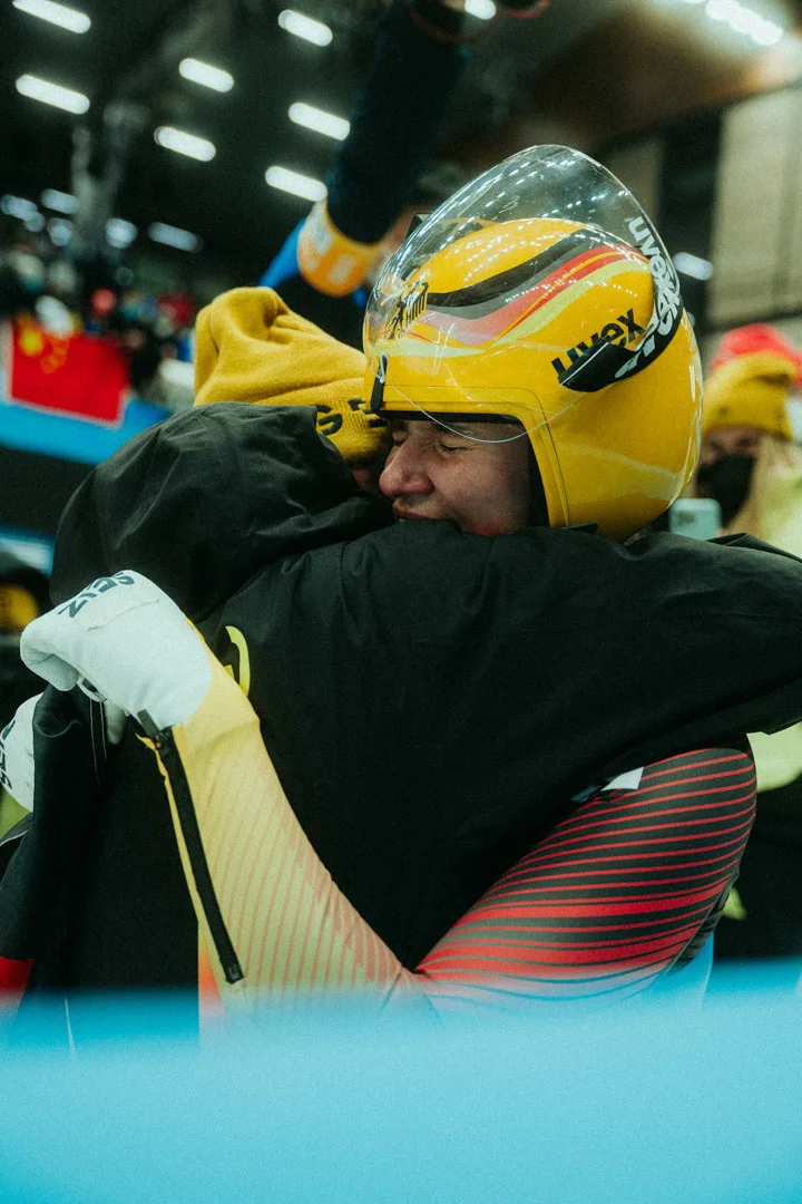A poignant close-up of two athletes sharing a tight hug after a competition. One athlete wears a yellow helmet with 