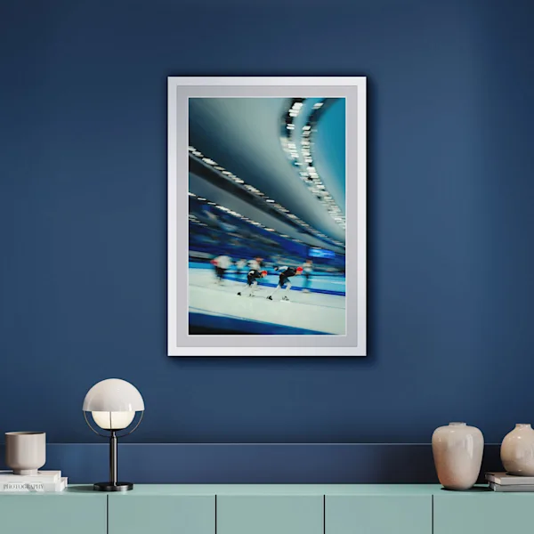 This winter sports photograph is presented in a natural white solid wood frame. The motif is elegantly bordered by a light grey mount. The image captures two speed skaters in action, set against a deep blue wall.