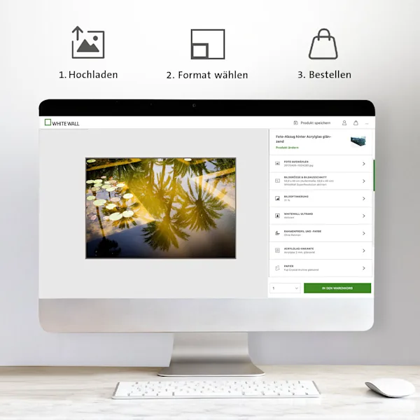 Ein silberner Desktop-Monitor auf einem hellen Holztisch zeigt den WhiteWall Produkt-Konfigurator. Zu sehen ist die Bearbeitung eines Foto-Abzugs hinter Acrylglas mit einem Motiv von Palmen-Spiegelungen. Über dem Monitor illustrieren drei Icons die Schritte: 1. Hochladen, 2. Format wählen, 3. Bestellen.