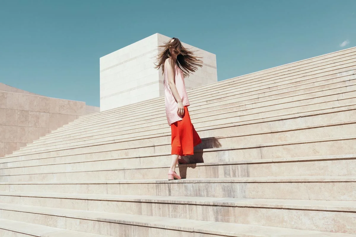 A woman in a red skirt and pink top stands on a wide stone staircase under a clear sky.