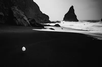 Black and white photo of a beach with dark sand, rocks, sea and a single balloon in the foreground.