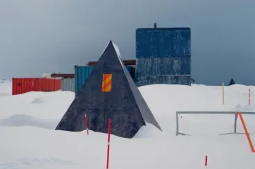 Estructura piramidal oscura, contenedores y edificios rectangulares se alzan en un paisaje nevado con postes indicadores.