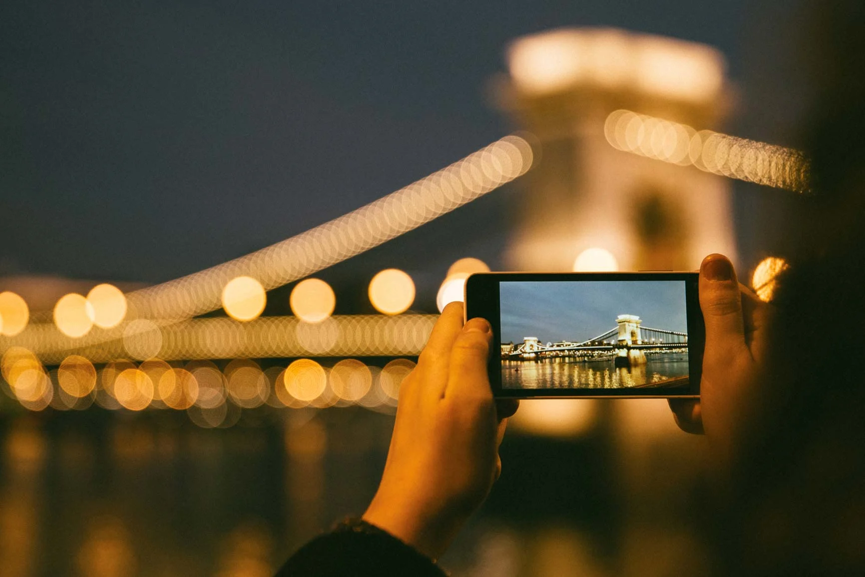 Persona fotografiando un puente colgante iluminado por la noche con un smartphone.