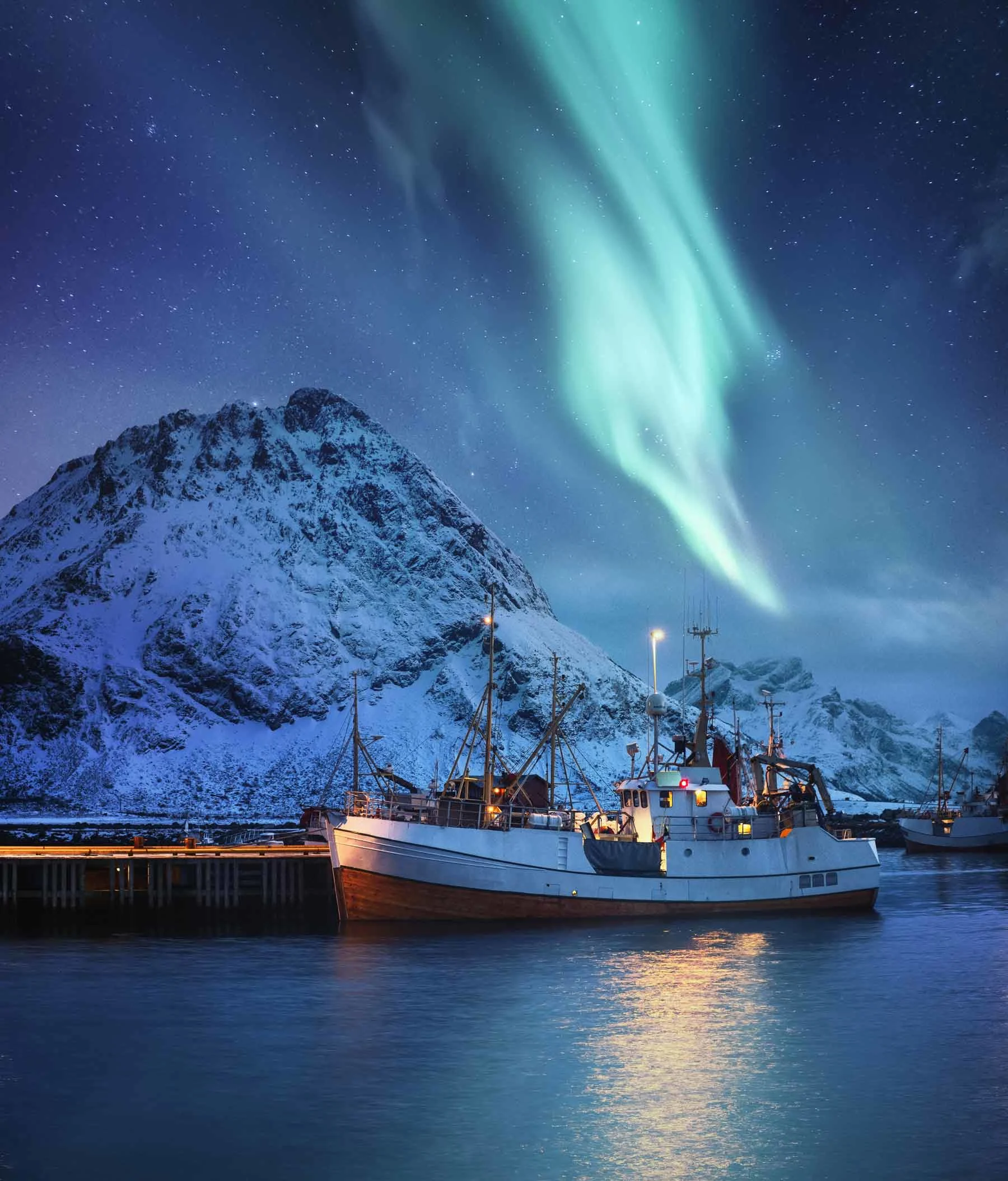 Fishing boat in harbor in front of snow-covered mountain under green northern lights in the night sky.