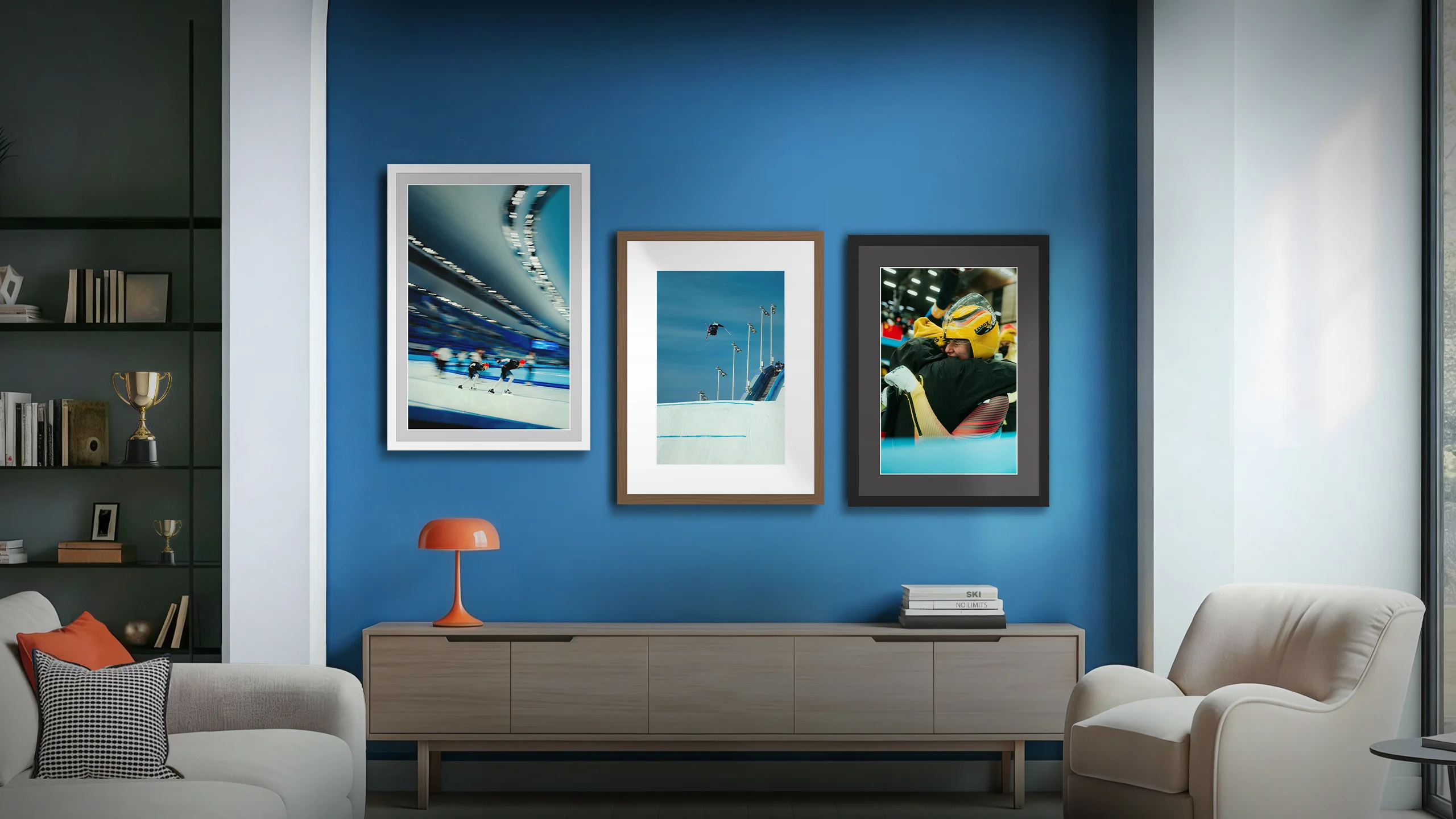 A modern living room interior featuring a blue accent wall. Three large framed prints of winter sports—speed skating, ski jumping, and an emotional embrace between two athletes—hang above a wooden sideboard. An orange lamp and some books are placed on the sideboard.