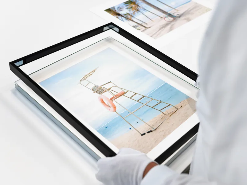 Person with gloves changes the frame of a magnet framed picture. The pictures shows a beach scene with a high seat for a lifeguard.