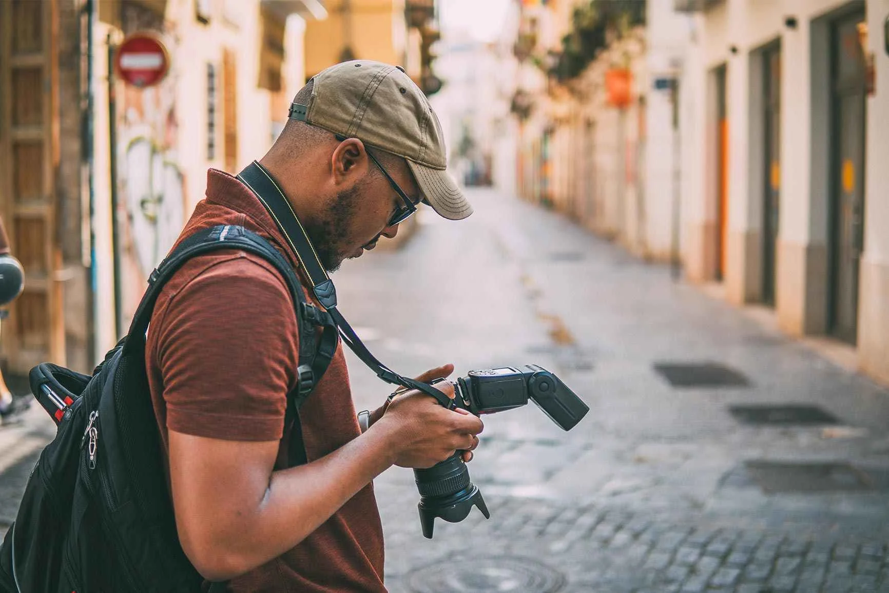 A man wearing a baseball cap and carrying a backpack is standing on a cobblestone street, holding a camera with a flash.