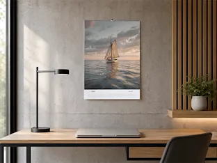 Un calendario de pared con un velero en el mar cuelga sobre un moderno escritorio de madera con un portátil y una lámpara negra.