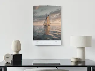 Modern desk setup with minimalist ceramic lamps, black clock, laptop, and wall calendar featuring sailboat at sunset