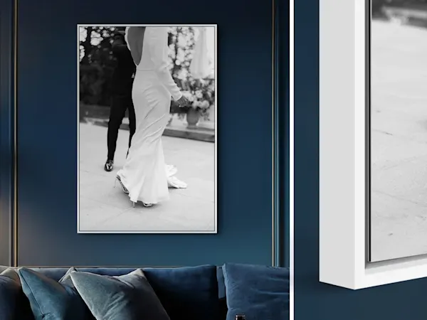 Une vue en deux parties montrant, à gauche, une photographie de mariage en noir et blanc grand format dans un élégant cadre caisse américaine blanc sur un mur bleu foncé. Le motif capture une mariée de dos dans une robe à dos nu et des talons hauts sur un sol en pierre, avec un marié debout derrière elle. En dessous, un canapé en velours bleu est visible. À droite, un gros plan détaillé met en valeur la profondeur et l'espace précis du joint de l'ombre du cadre en bois blanc.