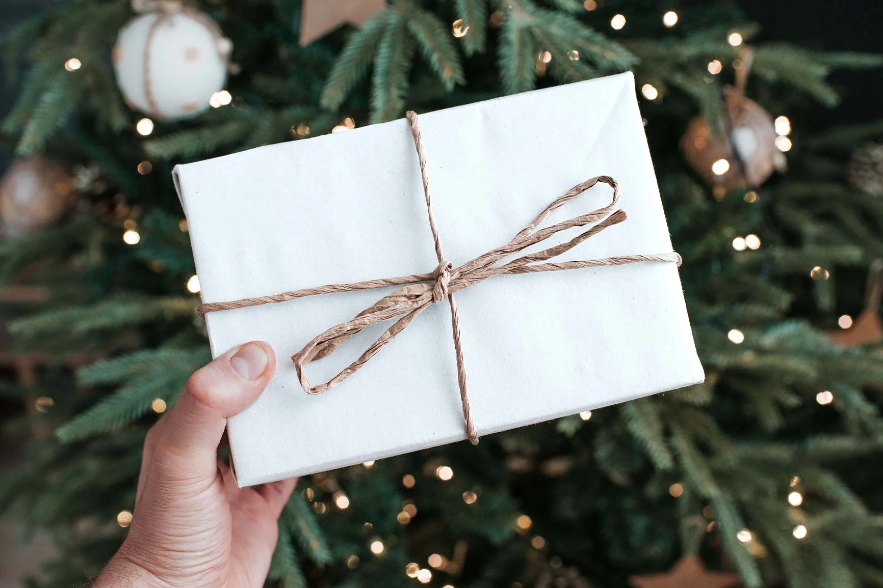 Weiße Geschenkbox mit Bindfaden umwickelt, die vor einem Weihnachtsbaum mit funkelnden Lichtern im Hintergrund gehalten wird.