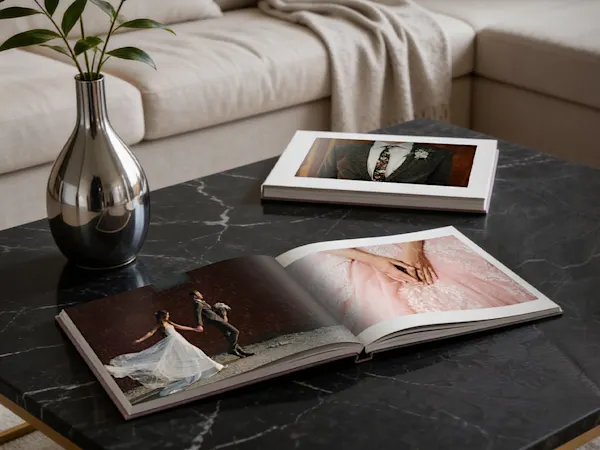 Two open wedding photo books on a dark marble coffee table beside a silver vase, with a beige sofa in the background.