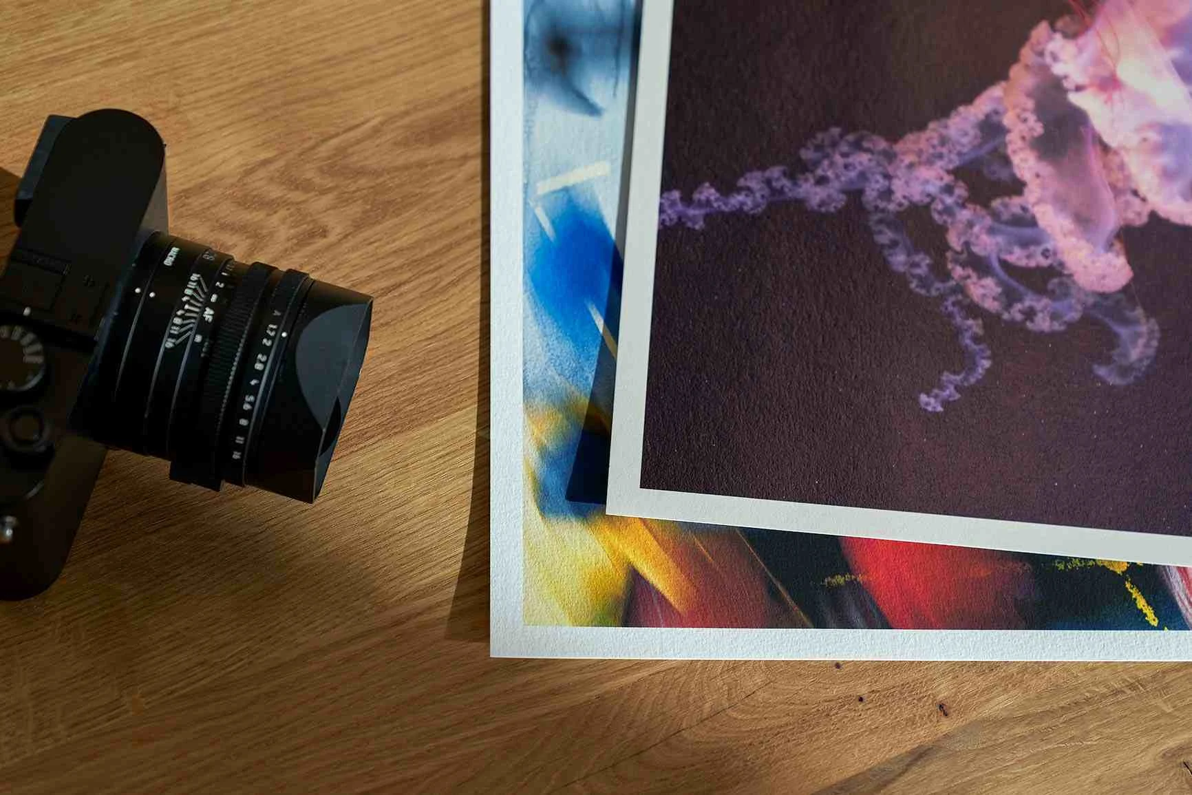 Zwei Fine Art Prints mit abstrakten, farbigen Mustern liegen auf einem Holztisch neben einem Kameraobjektiv.